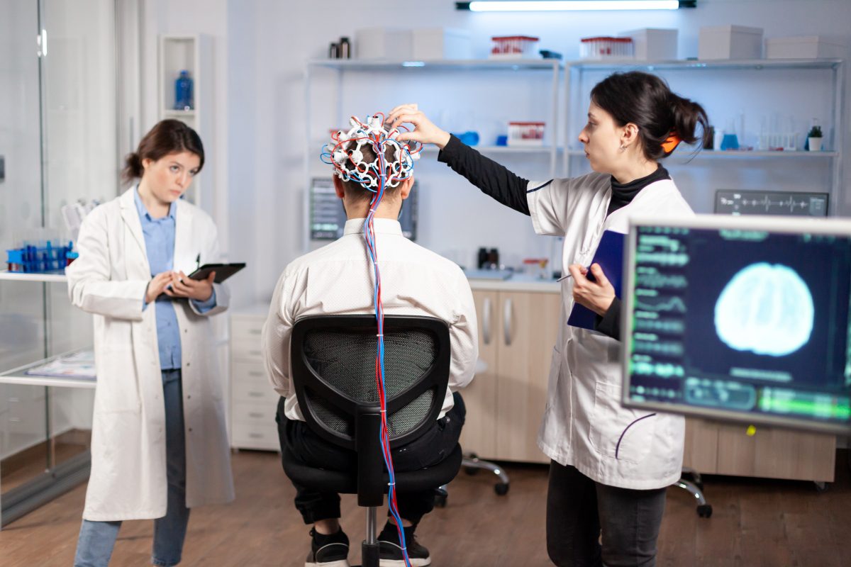back-view-man-patient-wearing-performant-brainwave-scanning-headset-sitting-neurological-research-laboratory-while-medical-researcher-adjusting-it-examining-nervous-system-typing-tablet